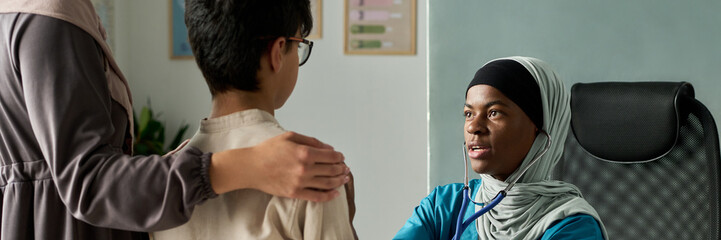 Muslim doctor in hijab attentively engaging with young boy seated beside parent, fostering...