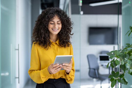 Latin American businesswoman in office using tablet, representing professionalism. Smiling in yellow blouse, standing in hallway with digital device, symbolizing technology in workplace. - Powered by Adobe