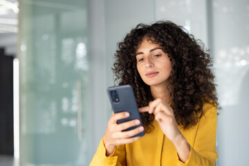 Latin American businesswoman with curly hair using phone in office. She wears yellow clothing,...