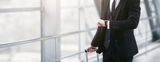 Cropped view of unrecognizable guy holding suitcase handle and checking time on wristwatch standing at airport