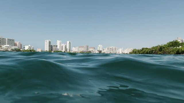 A scenic view of a coastal city skyline features tall buildings and calm ocean waters in the foreground at Condado Lagoon, Puerto Rico, under a clear blue sky.