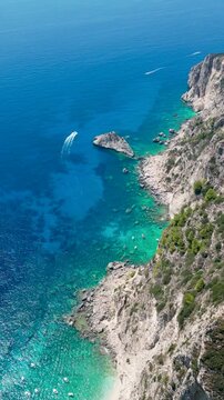 Aerial view of turquoise sea and blue, sparkling water in Palaiokastritsa, Corfu, Greece, from Angelokastro castle. Rocky coastline, three boats cruising, one racing. Vertical video, drone shot.