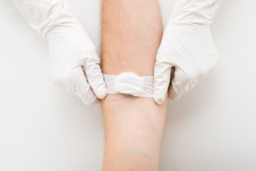 Doctor hands in white protective rubber gloves putting bandage with cotton wool on young adult man arm vein after blood test on light gray table background. Closeup. Patient first aid. Top down view.