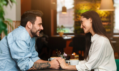 Lovely Couple In Love Flirting And Talking Holding Hands Having Date Sitting In Cozy Cafe. Romantic Relatioship. Side View