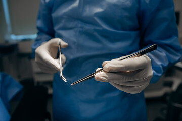 Close-up photo of doctor surgeon with clamp and forceps and hands in gloves and sterile suit under surgical lamp.