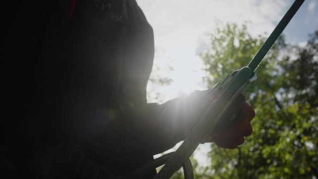 Climber illuminated by sunlight operates an ascender device, skillfully ascending a rope amidst trees. The scene captures the essence of outdoor adventure and athletic determination