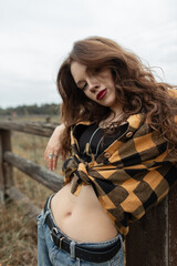 Beautiful fashion rural woman in fashionable autumn clothes with a Plaid Twill Western Shirt and vintage jeans sits near a wooden fence in the countryside