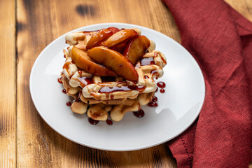 Belgian waffles with apples stewed in wine sauce on a white plate on a wooden background with a burgundy napkin.