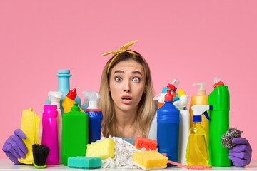 Shocked millennial woman holds cleaning supplies on pink background, free space