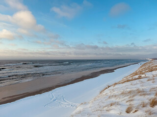The image shows a snowy beach with a clear view of the sea and the sky. The beach is covered in snow except for some areas near the water, and there are small waves in the sea under a partly cloudy sk