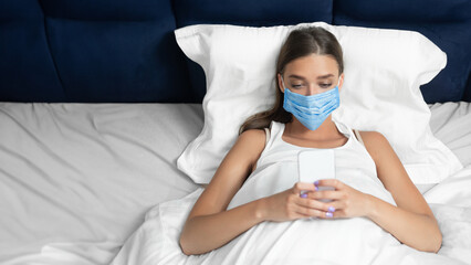 Lady In Protective Medical Mask Using Cellphone Application Lying In Bed At Home During Coronavirus...