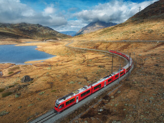 Aerial view of modern red train, river, alpine mountains, orange hills and trees, sky with clouds...