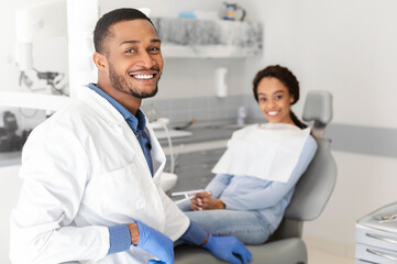 Obraz premium Visiting dentist. Black dentist doctor and female patient in chair smiling at camera, clinic interior