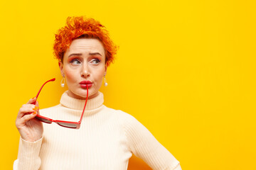 young red-haired uncertain woman with curly hair holds sunglasses and thinks on a yellow isolated...