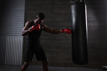 Fototapeta premium young strong african american boxer in red gloves hits punching bag, muscular athlete trains punch in dark gym and does sports on black background