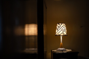 A close-up of a small table lamp on a nightstand in a dimly lit room. The lamp has a patterned fabric shade and casts a warm glow. The rest of the room is dark, with only the soft light from the lamp 