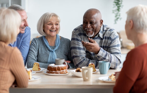 International group of positive men and women sitting around table drinking tea with cake and having conversation, smiling and laughing, having home party or enjoying time at nursing home