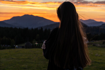 A woman with long hair stands in a field, looking at the sunset over a mountainous landscape, creating a peaceful silhouette.