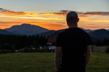 Fototapeta premium A man stands with his back to the camera, gazing at the sunset over mountains in a peaceful rural setting, creating a contemplative mood.