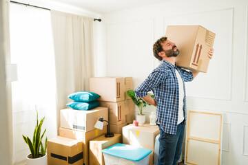 Struggling man holding his back while lifting heavy cardboard box during move into new apartment