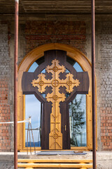 A close-up of a wooden Orthodox cross door under construction, set against an unfinished brick wall, showcasing intricate carvings and craftsmanship.