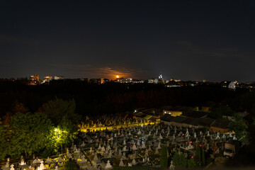 A nighttime urban landscape featuring a cemetery illuminated by soft lights, with city buildings in the background and the moon rising on the horizon.