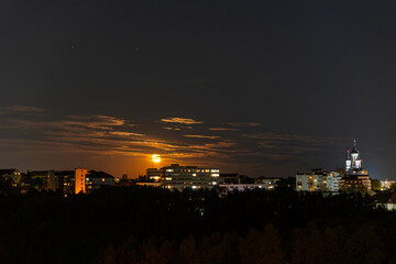 A nighttime cityscape featuring buildings with illuminated windows and the moon rising amidst clouds, creating a serene and atmospheric scene.