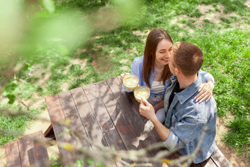young married couple in denim clothes sitting in the garden at wooden table and drinking coffee and kissing on date in the summer, girl and guy having breakfast in outdoor cafe and flirting