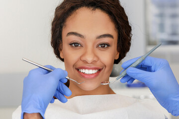 Portrait of smiling african lady and cropped dentist hands in blue gloves with tools in