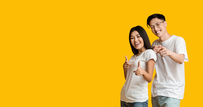 Hey there. Friendly asian man and yoman indicating at camera over yellow background, copy space