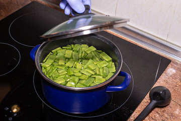 Man's hand with nitrile glove uncovering a pot containing flat green bean pot containing green flat...