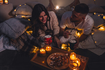 Cozy romantic evening, home weekend In bed for a loving couple. Candles, movies, hot chocolate with marshmallow, popcorn. Beautiful young man and woman enjoying relationship, having fun, laughing