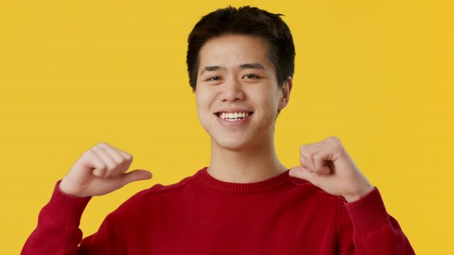 A cheerful young Asian man wearing a red sweater smiles broadly while pointing to himself, radiating confidence and happiness in front of a vibrant yellow backdrop.