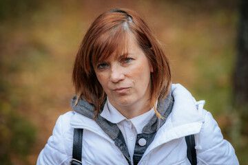 A close-up portrait of a woman with auburn hair wearing a white jacket, standing outdoors. She has...