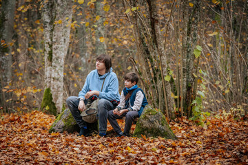 A young adult and a child sit on a bench surrounded by fallen leaves in an autumn forest. One looks up while the other gazes forward, enjoying the peaceful surroundings