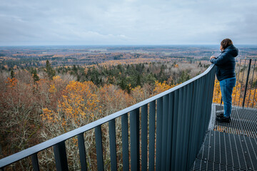 A person stands on an observation deck, looking out at a vast autumn forest with vibrant orange, yellow, and green trees. The landscape stretches out under a cloudy sky. © Emvats