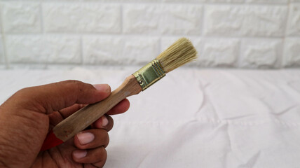 Hand holding a small paintbrush with a wooden handle and bristle tip, against a white brick wall...