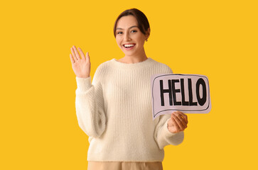 Beautiful woman holding paper speech bubble with word HELLO and waving hand on yellow background © Pixel-Shot