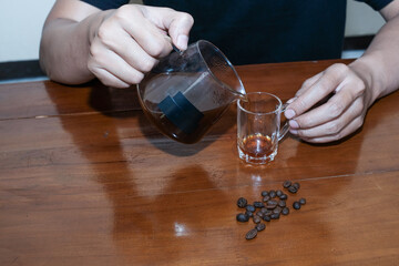 Closeup of a hand pouring drip coffee into a small glass on wooden table, making coffee homemade.