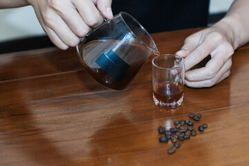 Closeup of a hand pouring drip coffee into a small glass on wooden table, making coffee homemade.