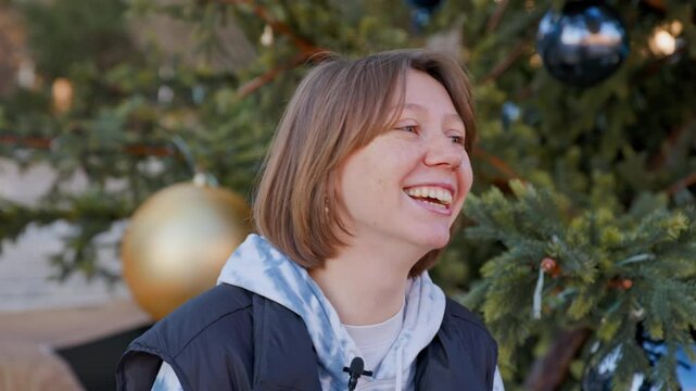 A smiling woman with a bob haircut stands cheerfully in front of a decorated christmas tree outdoors.