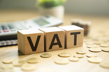 Wooden blocks spelling VAT placed on a desk with coins, calculator, and notebook, representing value-added tax, financial calculations, business taxation, and economic policy