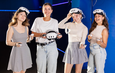 Four young female friends equipped with VR goggles and game controllers, thrilled after exciting...