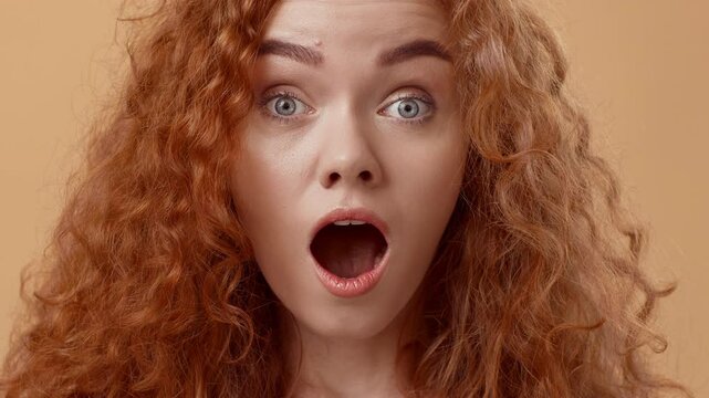 A young woman with vibrant curly red hair shows a surprised expression, her mouth open and eyes wide. The warm beige background complements her expressive demeanor.