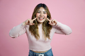 A cheerful young asian woman is smiling and making a playful gesture with her fingers. She has long, wavy hair. Portrait on a simple pink background with studio light.