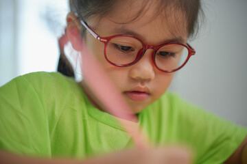 Fototapeta premium Asian young girl wearing glasses is writing with a red pencil. She is focused on her work and she is in a serious mood