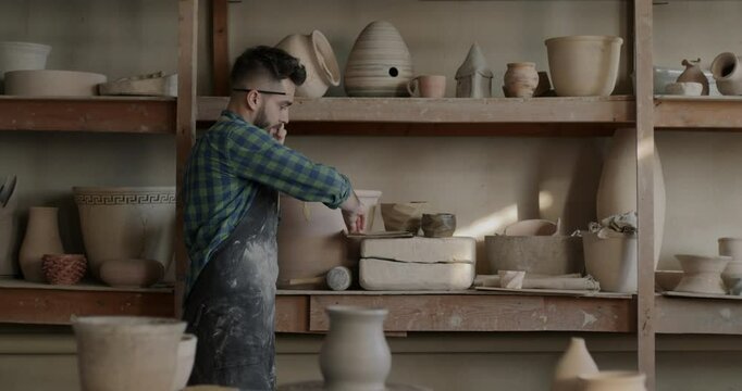 Small business owner talking on mobile phone checking pots on shelves developing pottery workshop. Communication and traditional arts studio concept.