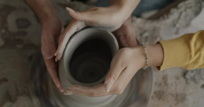 Close-up of male and female hands making pot together while couple enjoying pottery class. People using throwing wheel working with clay.