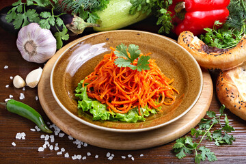 Korean carrot salad in a plate against the background of fresh vegetables, on a wooden board. Cafe, lunch, close-up.