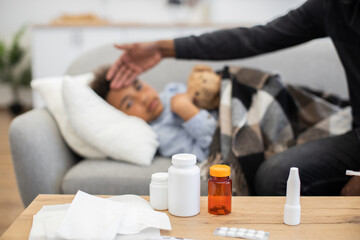 Caring father checks temperature of sick African boy laying on couch. Child is wrapped in blanket,...